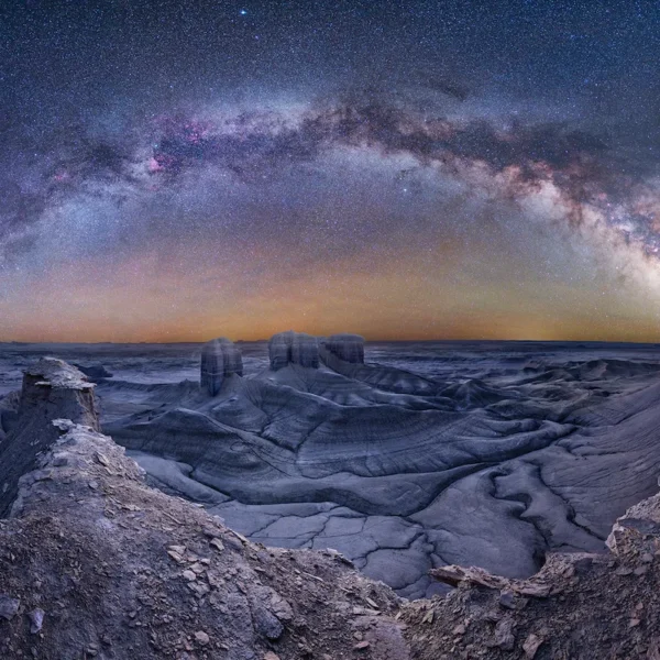 A panoramic night‑sky photograph showing the Milky Way arching above the barren, lunar-like landscape of Moonscape Overlook in Utah, captured from a vantage point.