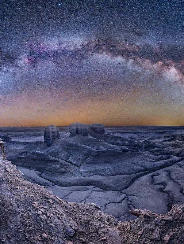 A panoramic night‑sky photograph showing the Milky Way arching above the barren, lunar-like landscape of Moonscape Overlook in Utah, captured from a vantage point.