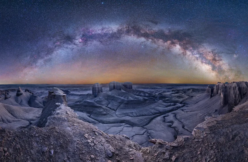A panoramic night‑sky photograph showing the Milky Way arching above the barren, lunar-like landscape of Moonscape Overlook in Utah, captured from a vantage point.