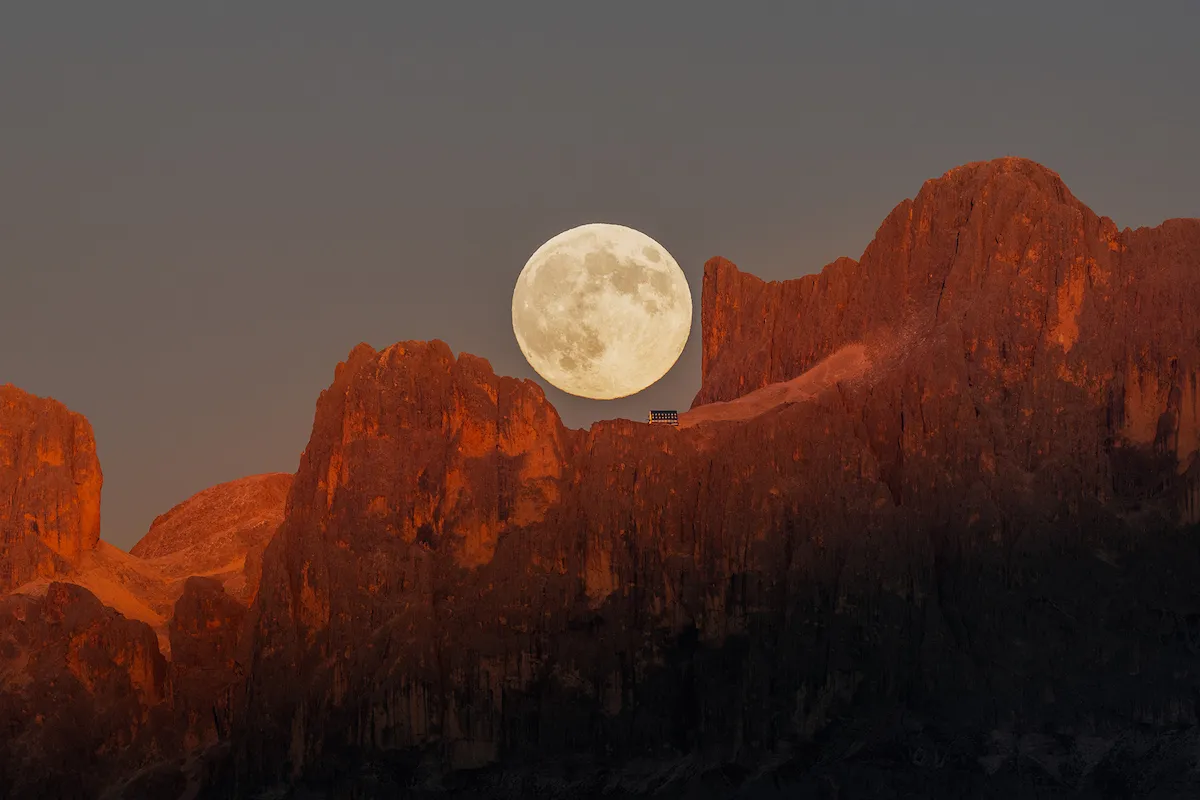 A photograph of the full moon rising above the rugged peaks of the Dolomites in South Tyrol with clear skies and the mountain tops catching the last light of day