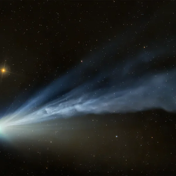 An image showing Comet 12P/Pons‑Brooks with a flowing, wind‑shaped tail, its blue tones contrasting with the warm glow of a nearby star.