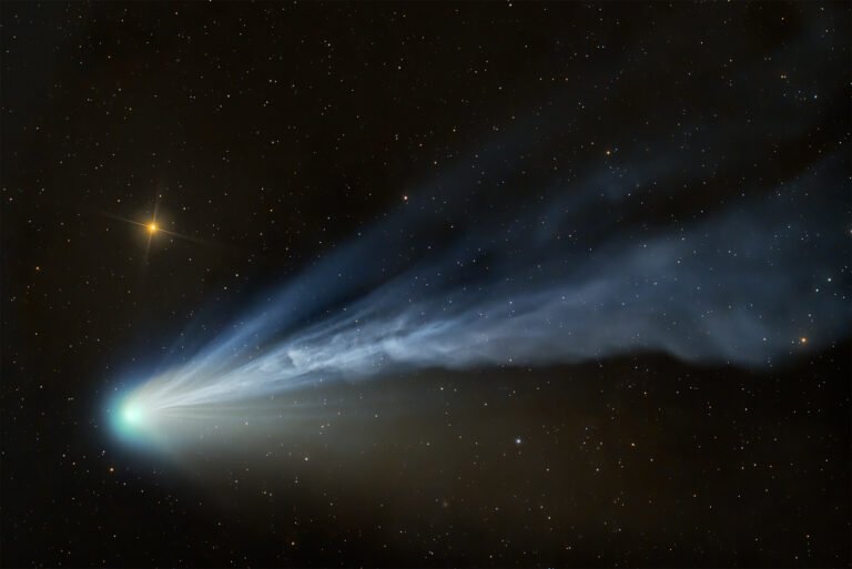 Comet 12P/Pons-Brooks with a wafting tail illuminated by solar winds, glowing blue against a golden starburst in the California night sky.