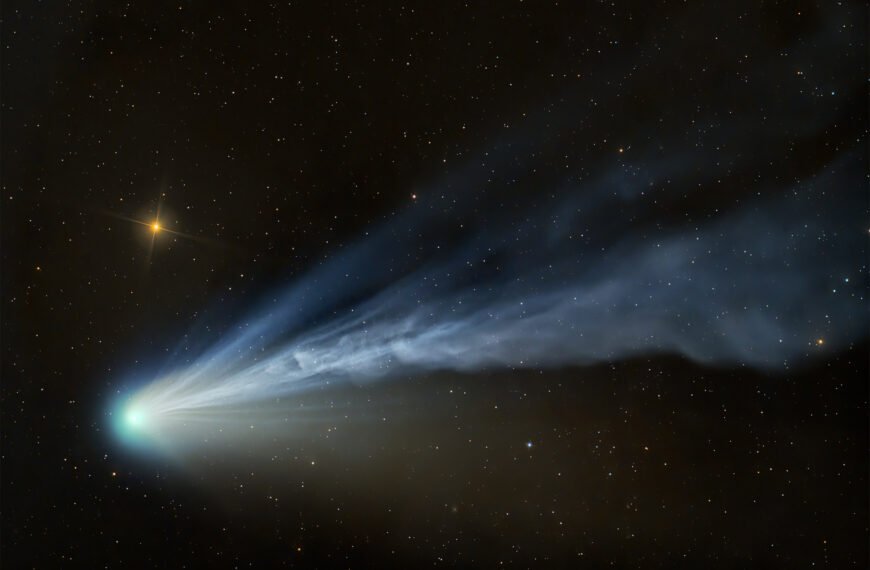 Comet 12P/Pons-Brooks with a wafting tail illuminated by solar winds, glowing blue against a golden starburst in the California night sky.